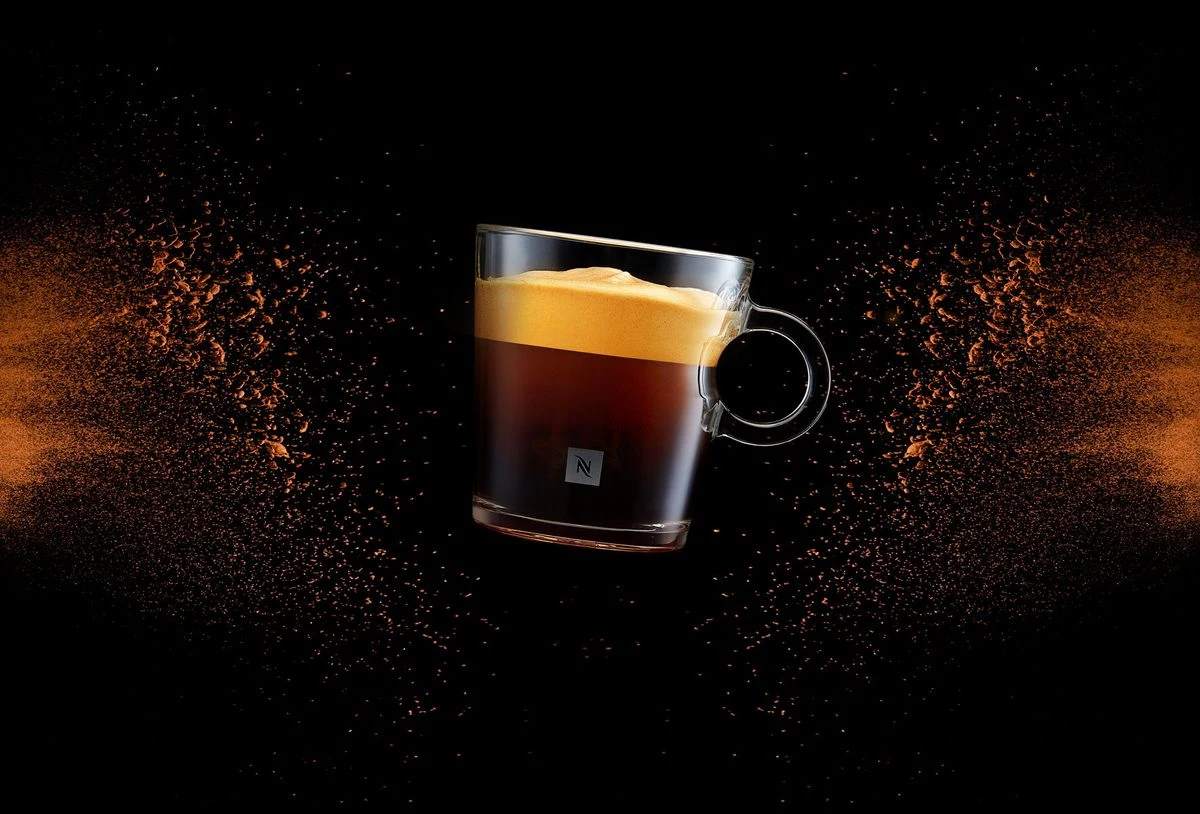 Une tasse de café Nespresso entourée de particules de café sur un fond noir.